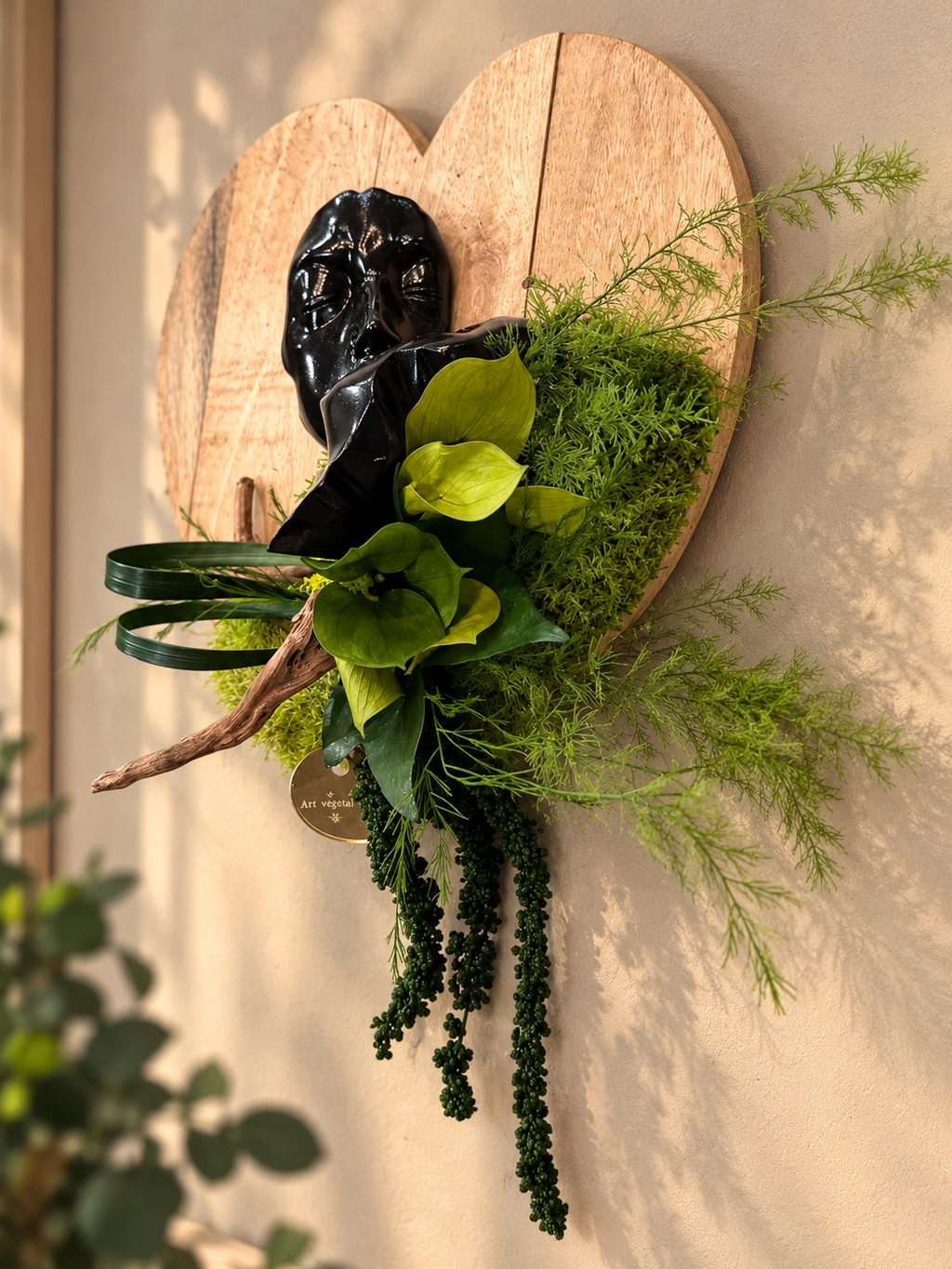 Décoration murale végétale artisanale sur support en bois naturel en forme de cœur, ornée d’une sculpture de visage noir, de feuillages verts stabilisés, de mousse et de branches décoratives\. Création originale et sans entretien, idéale pour une décoration intérieure naturelle et contemporaine\.